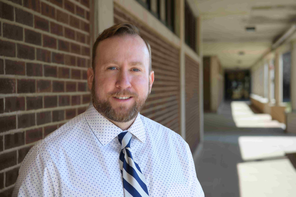 Derek Weber stands to the left of the image, wearing a white shirt with a striped blue tie.